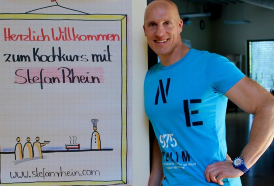 Stefan Rhein posiert neben einem Plakat, auf dem steht "Herzlich Willkommen zum Kochkurs mit Stefan Rhein"